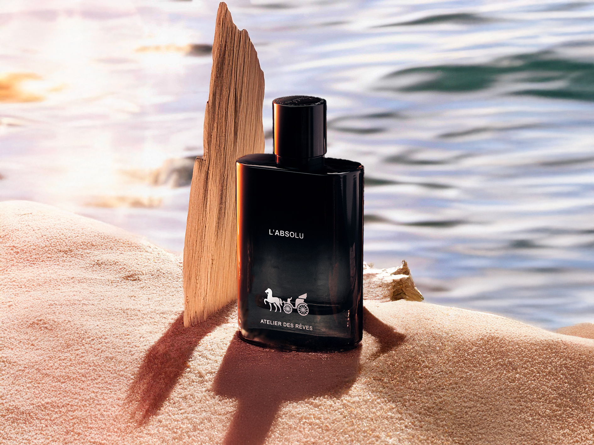 Perfume bottle labeled 'L'Absolu' on a sand with a piece of oud wood and water  in the background